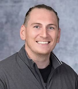 Headshot of Tim Hazen. He is a Caucasian man with brown hair tied back in a ponytail. He is wearing a grey quarter-zip jacket with a black t-shirt on underneath 